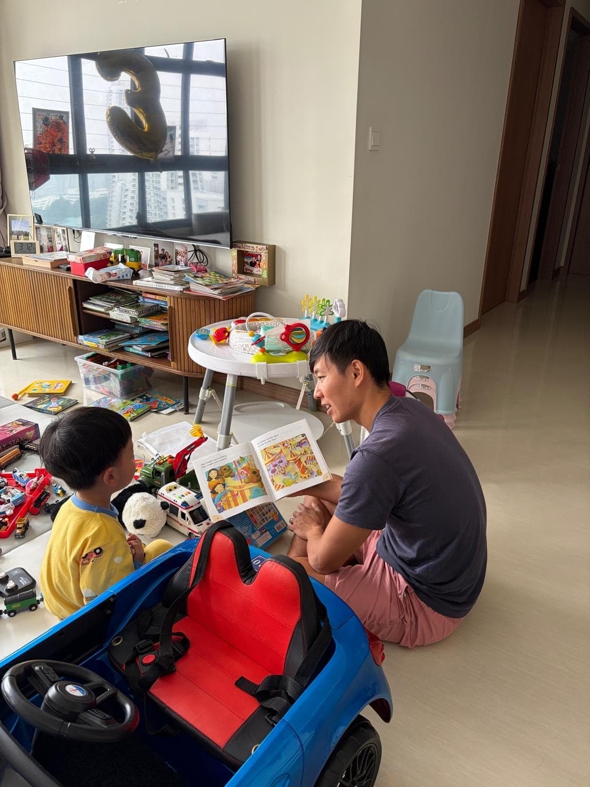 Alfred reading a picture book to his son on the living room floor, surrounded by toys